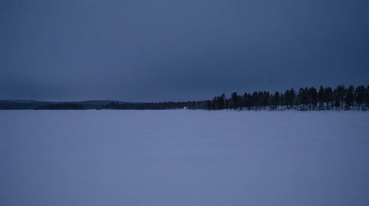 A vast frozen lake covered in smooth snow stretches toward a low line of dark forest under a pale, starless sky.