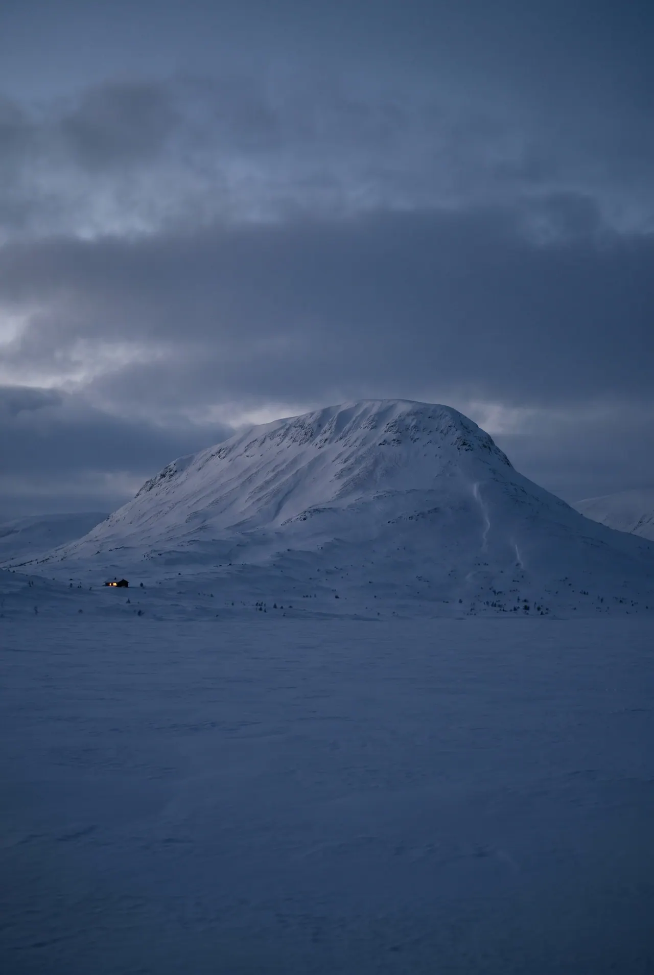 The steep rounded flank of a fell rising from a snowfield, its ridges traced in wind-blown snow beneath a dim sky.
