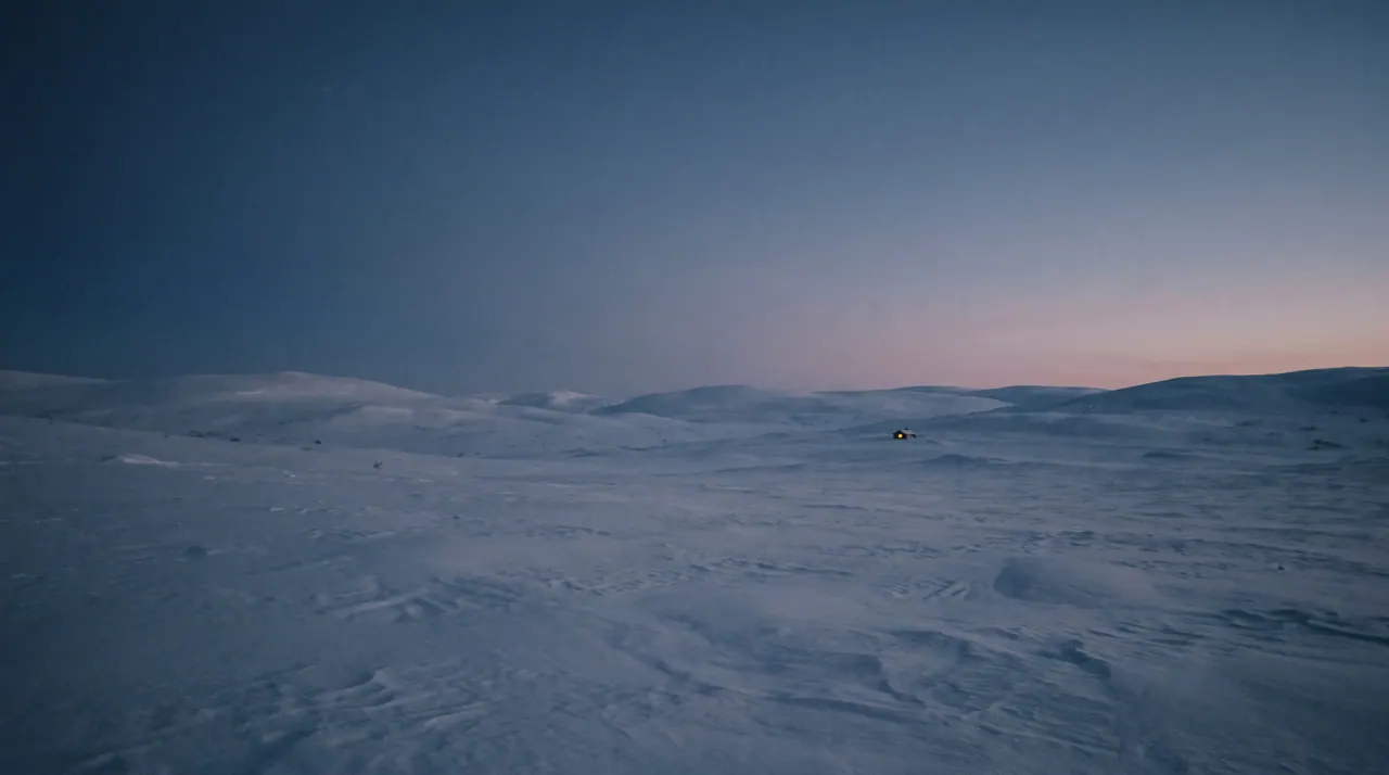 A wide winter landscape at midday during the polar night: rolling snow-covered hills under a sky that brightens only faintly near the horizon.
