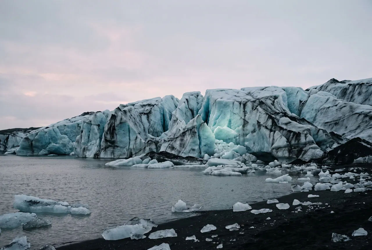 The fractured face of a glacier rising out of grey water, its deep crevasses streaked with dark ash and pale turquoise light.