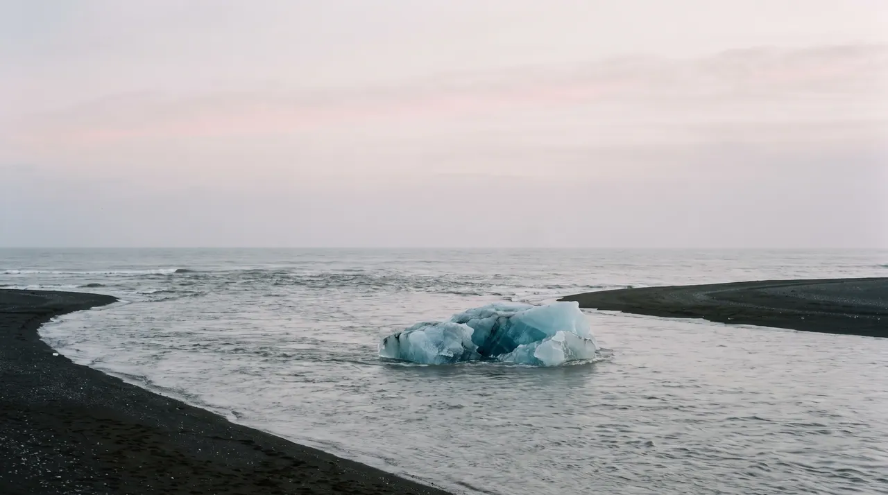 A lone iceberg drifting out through the mouth of the lagoon toward an open grey ocean, with a narrow strip of black beach on either side.